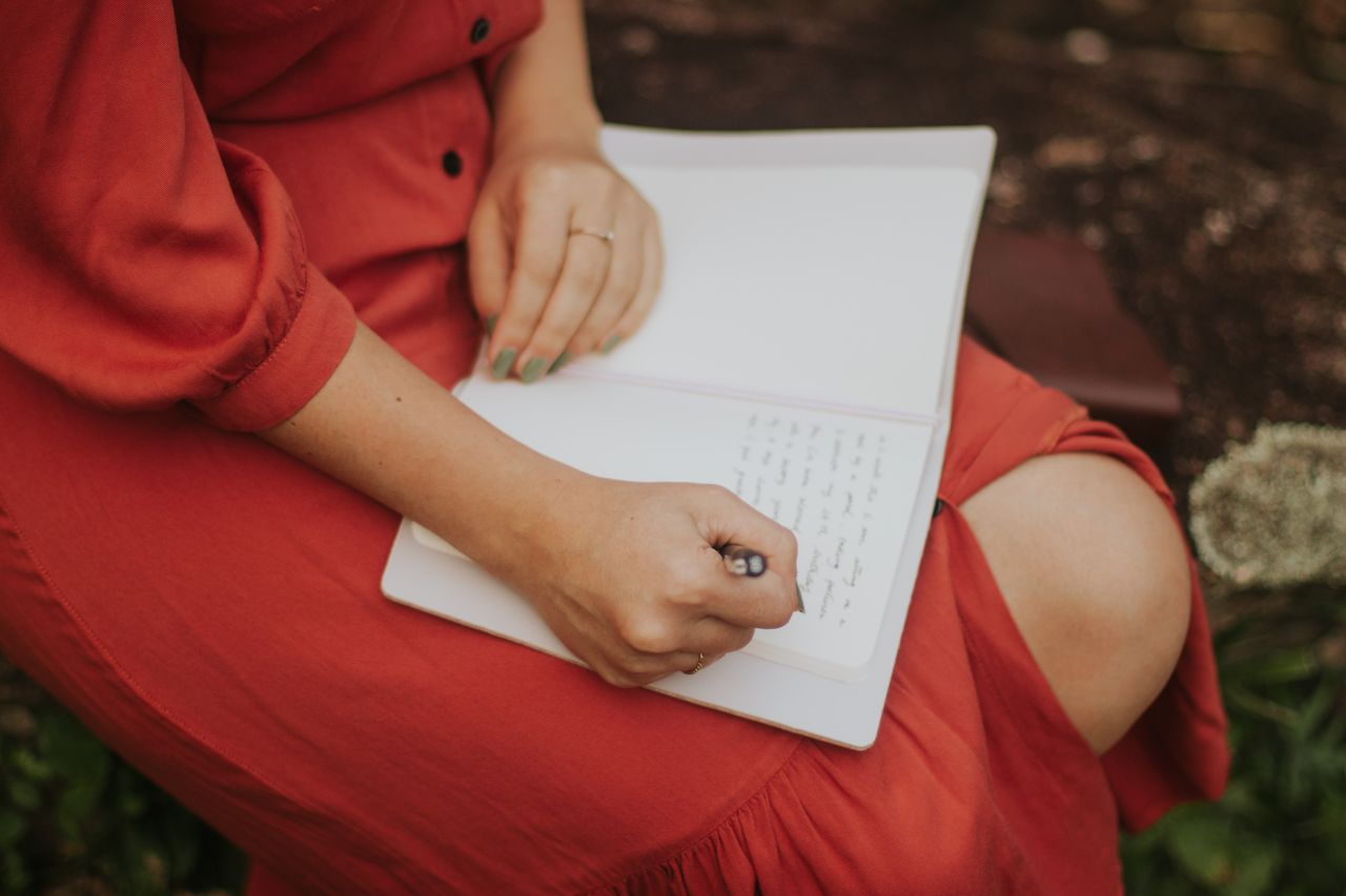 services-04 A woman in a red dress writing in a journal, outdoors over a leafy background.